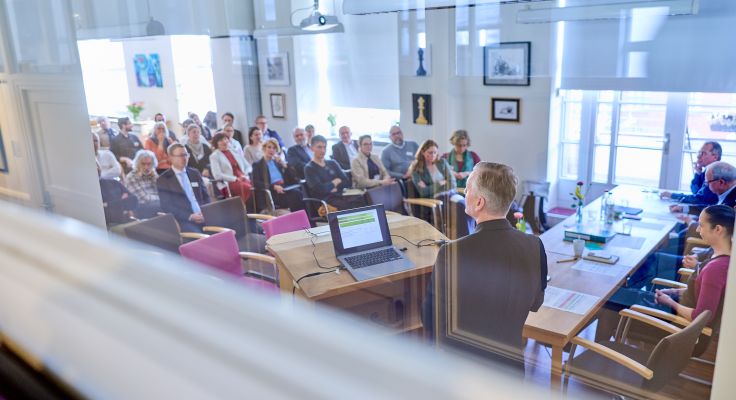Gäste sitzen in Reihen im Tagungsraum in Richtung des Vortragenden  