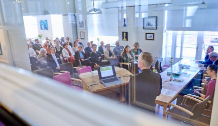 Gäste sitzen in Reihen im Tagungsraum in Richtung des Vortragenden  