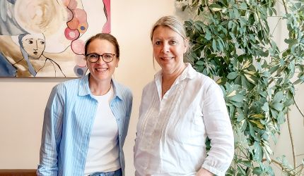 Miriam Jorke und Britta Buchholz stehen vor Kunstbild und Grünpflanze
