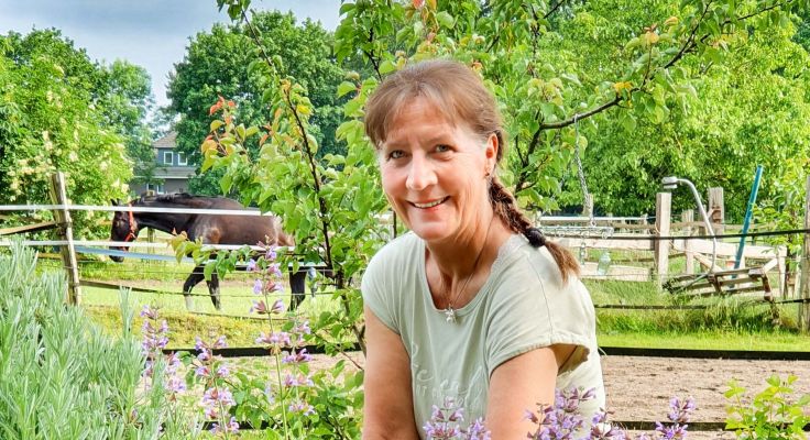 Petra Voltmer sitzt entspannt in einem Garten mit einem stehenden Pferd im Hintergrund 